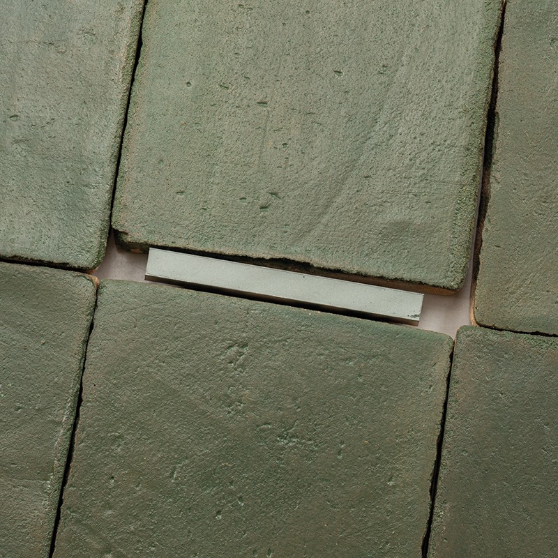 Matte groene keramische tegel met licht oneffen oppervlak; omliggende identieke tegels en een smalle, lichtgekleurde voeg met zichtbare witte strip ertussen.