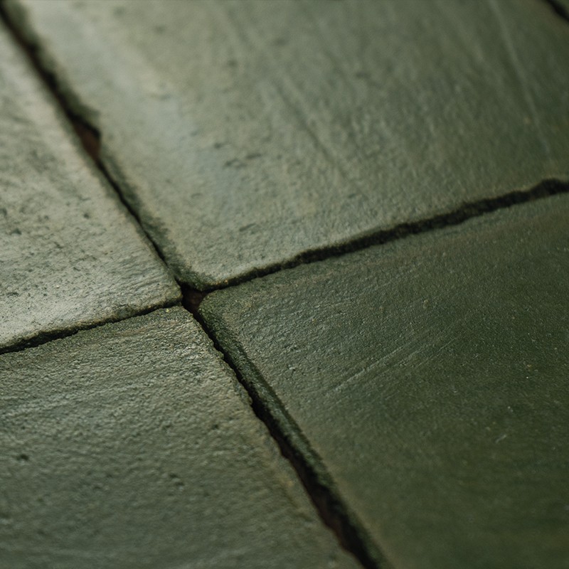 Azulejo verde mate, bordeado por juntas oscuras formando una cruz; superficie rugosa con ligero brillo, adyacente a otros azulejos similares en un plano inclinado.