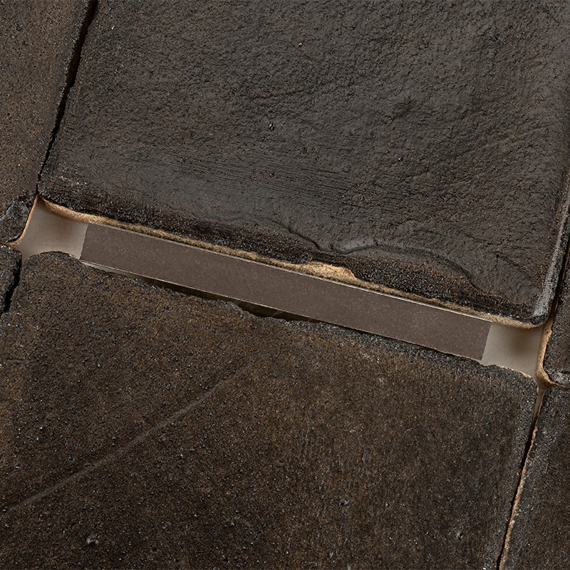 Azulejo oscuro rectangular, borde superior desgastado; junta hundida con tira metálica visible; entorno: otros azulejos rugosos y suciedad acumulada en las juntas.