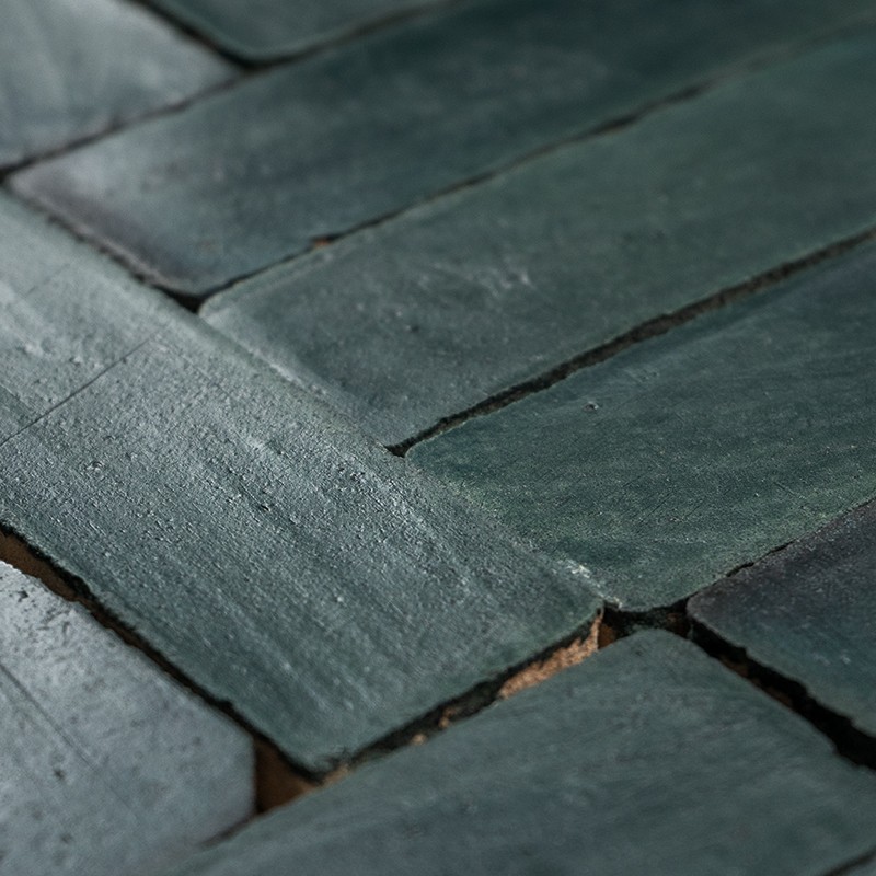 Azulejos retangulares verde-escuro, superfície esmaltada levemente irregular, assentados em padrão escalonado; rejunte escuro e pequenas lascas expostas entre as peças.