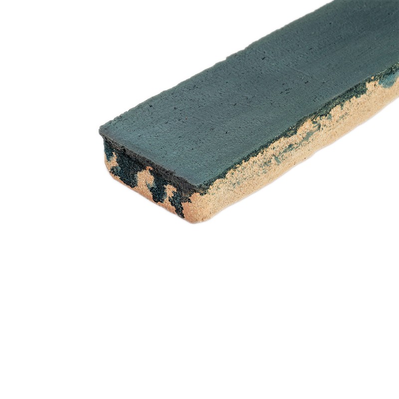 Smalle keramische tegelstrip met donkergroene bovenlaag en beige poreuze onderkant, afgesleten randen; ligt los op witte achtergrond.