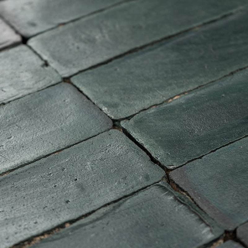 Azulejos rectangulares verde oscuro con superficie envejecida y pequeñas grietas; juntas oscuras poco uniformes, dispuestos en filas inclinadas sobre soporte rígido con luz lateral suave.