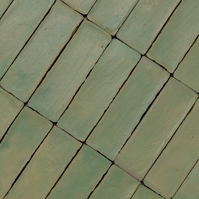 Azulejos rectangulares verdes, superficie mate y desgastada con variaciones de tono; dispuestos en orientación diagonal, separados por juntas oscuras e irregulares que acentúan los bordes.