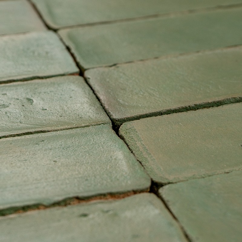 Carrelage rectangulaire vert pâle; joints foncés visibles, surfaces légèrement irrégulières et brillantes, usure aux bords; plan rapproché pris en oblique.