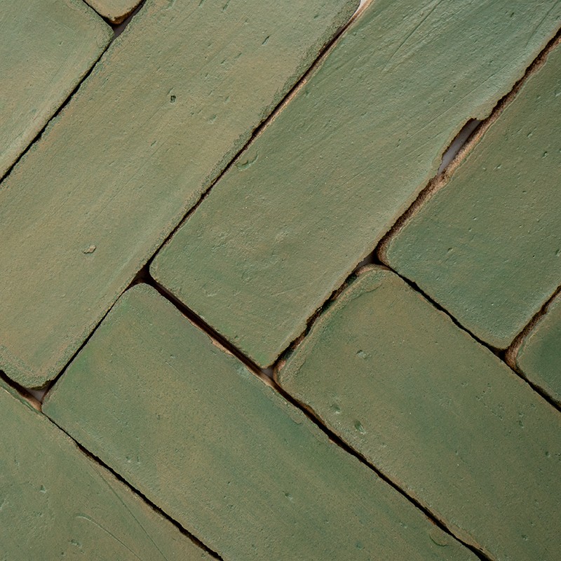 Groene rechthoekige keramische tegels met licht afgeronde randen, geplaatst in visgraatpatroon; duidelijke voegen, lichte oppervlakkige oneffenheden en kleurvariaties.