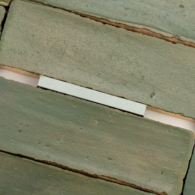 Langwerpige groene keramische tegel in verspringende horizontale rijen, ruwe textuur en dunne voeg; smalle witte plastic afstandhouder zichtbaar tussen twee tegels.