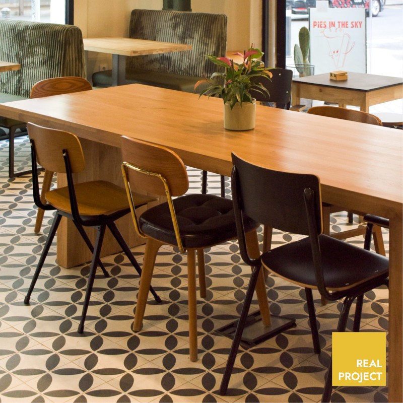 Azulejo geométrico circular en suelo; mesa larga de madera con sillas y planta, interior de cafetería junto a ventanal. Texto: "PIES IN THE SKY"; "REAL PROJECT".
