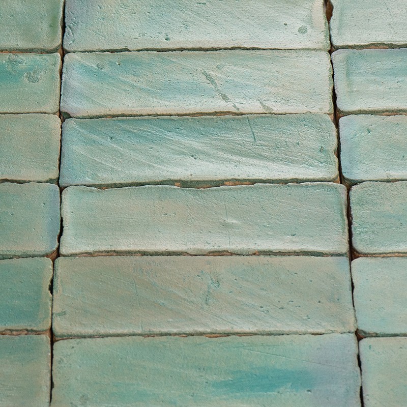 Carrelage rectangulaire émaillé bleu-vert, disposé en briques horizontales; joints visibles, surfaces légèrement irrégulières et écaillées, variations de teinte, fond indistinct.