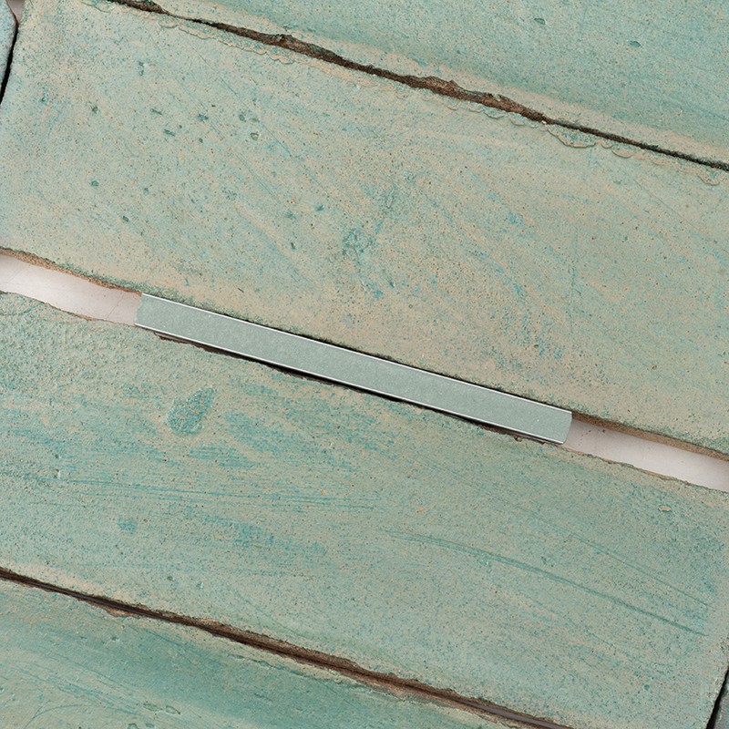 Carrelage turquoise usé à surface mouchetée, trois lattes horizontales séparées par joints clairs; petite pièce rectangulaire métallique posée transverse sur un joint.