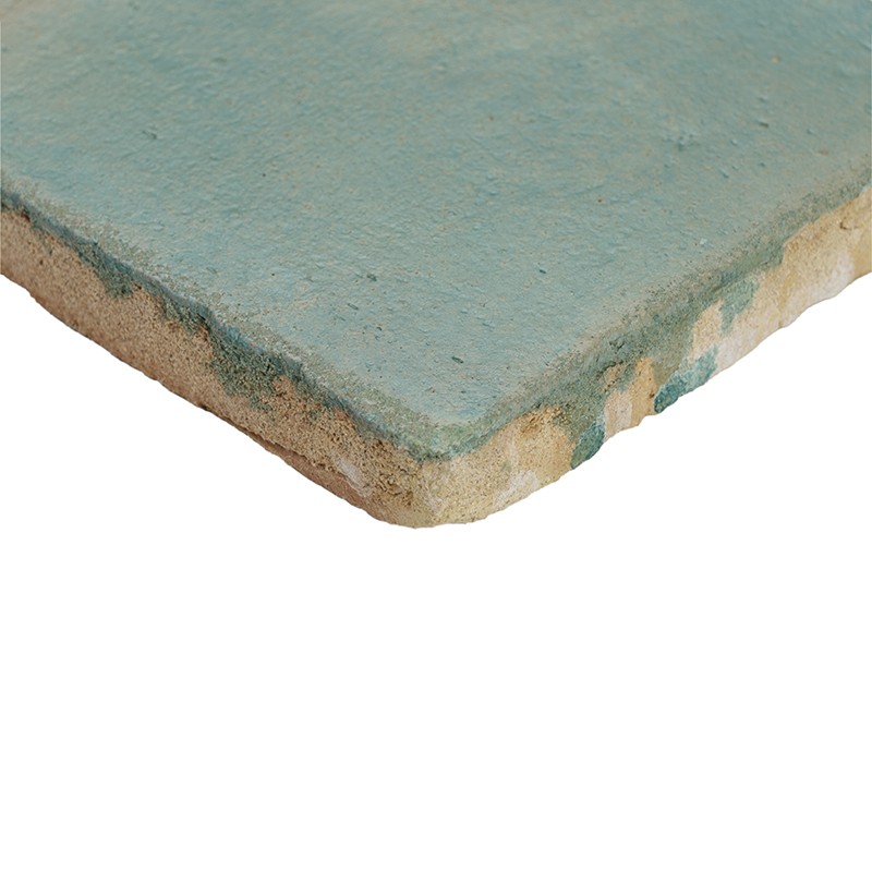 Carreau rectangulaire turquoise à bord usé révélant pâte beige, angle en avant-plan, surface légèrement texturée et écaillée, présenté sur fond blanc neutre.