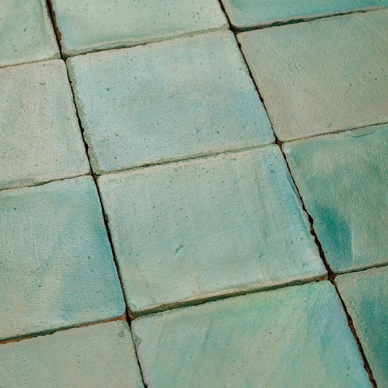 Azulejos cuadrados verde-azulados desgastados; lechada oscura entre ellos, superficie irregular y brillo mate, dispuestos en patrón de rejilla, posiblemente suelo o revestimiento.