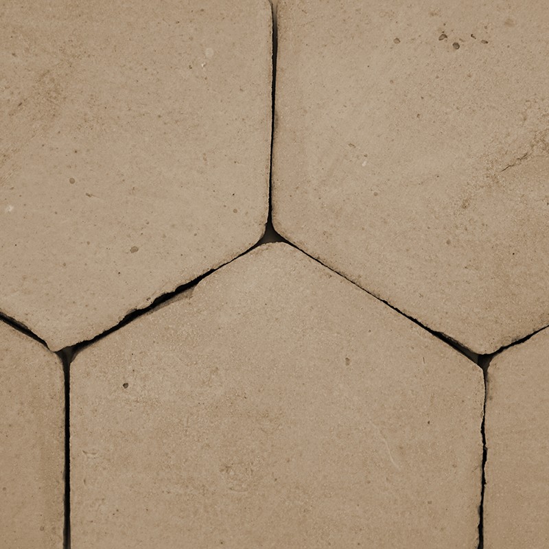 Beige hexagonal tile, rough surface with dark cracked stains and joints; pattern of adjacent tiles forming a continuous pavement.