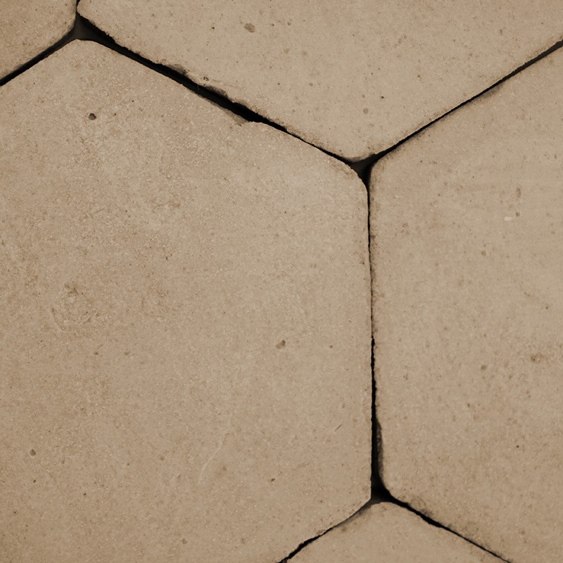 Beige hexagonal tile with narrow joints and visible cracks; slightly worn surface, alongside other tiles forming a continuous pavement.