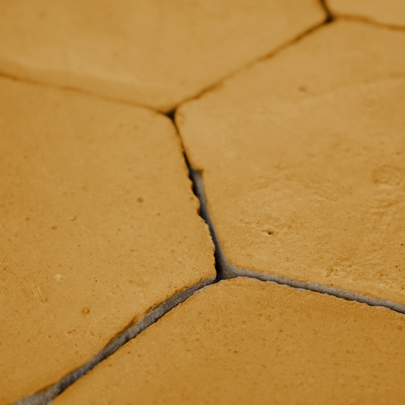Azulejo hexagonal amarelo envelhecido; juntas estreitas escuras, fissuras visíveis e superfície irregular, formando piso de peças justapostas.