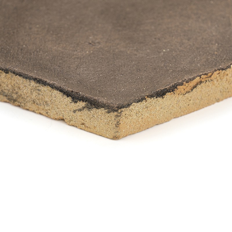 Carreau carré brun foncé vu en gros plan, bord inférieur montrant noyau beige granuleux et couche de surface sombre, fond blanc.