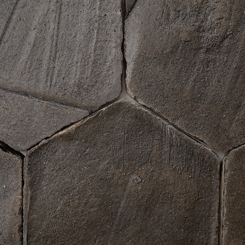 Azulejo hexagonal de pedra escura; juntas visíveis e superfície áspera e desgastada, com pequenos riscos e sujeira, em piso contínuo de peças iguais.