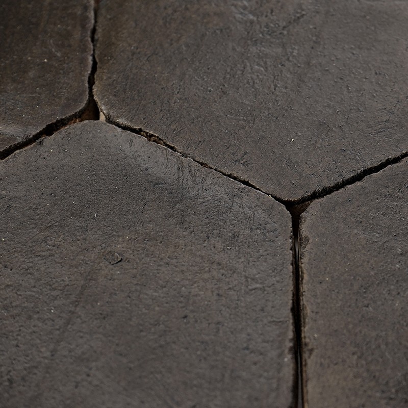 Carrelage hexagonal sombre, surfaces usées et légèrement irrégulières; joints profonds et fissurés, texture rugueuse visible sous un éclairage latéral faible.