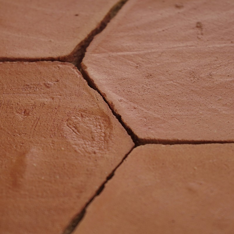 Carrelage hexagonal en terre cuite, couleur rouille; joints foncés et fissurés, surfaces légèrement usées avec minuscules empreintes et texture granuleuse.