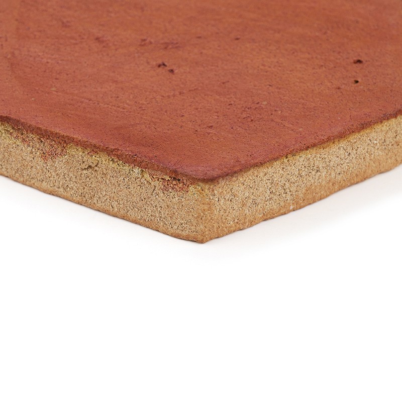 Carrelage en terre cuite: coin vu en gros plan, surface rougeâtre lisse, tranche inférieure rugueuse couleur sable, arête biseautée, fond blanc neutre.