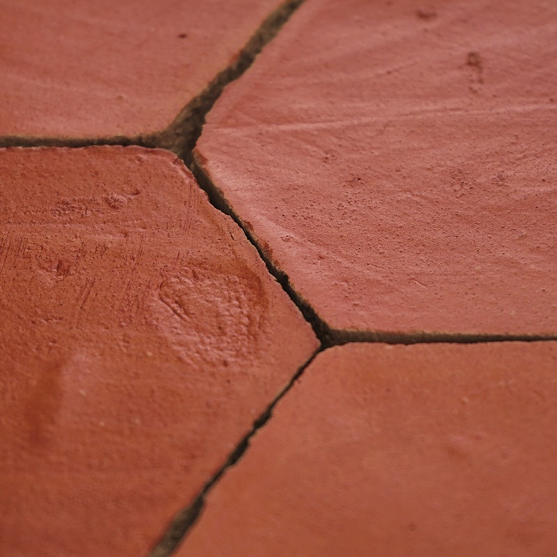 Carrelage hexagonal en terre cuite orangée, gros plan montrant joints foncés et fissures, surface mate légèrement irrégulière avec traces d'usure.