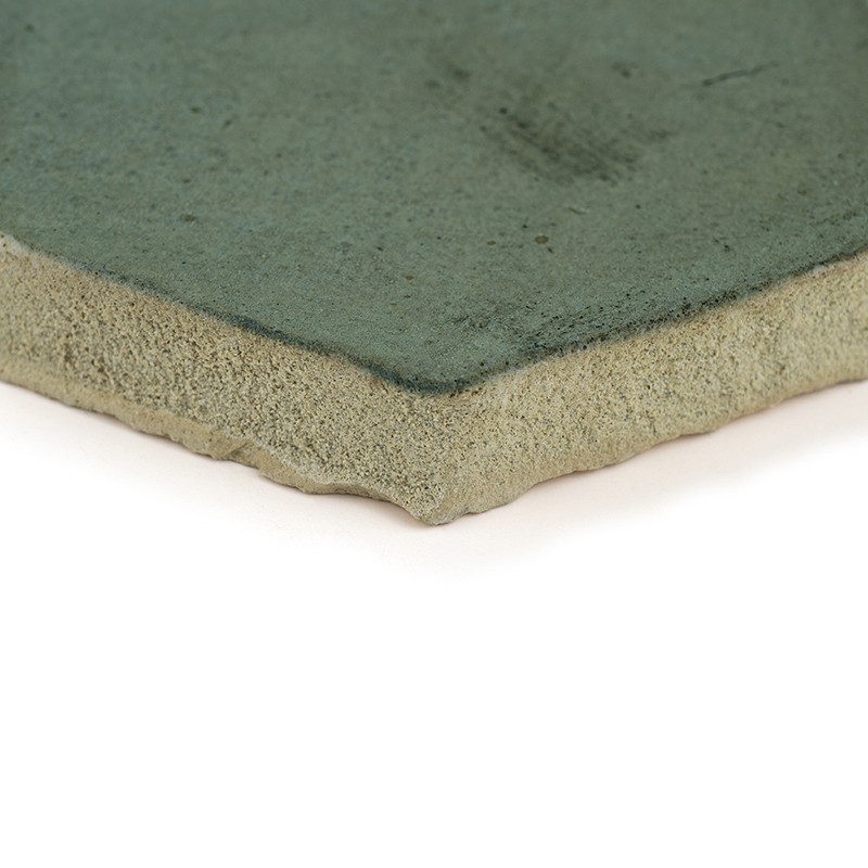 Azulejo verde: vista en primer plano de su esquina, superficie esmaltada verde y respaldo poroso beige, apoyado sobre fondo blanco iluminado.
