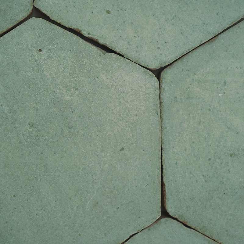 Azulejos hexagonales verdes desgastados; juntas estrechas oscuras y pequeñas grietas, superficie con manchas y textura rugosa formando un pavimento.