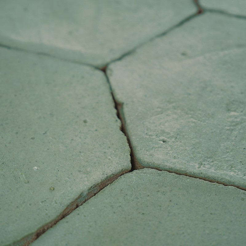 Azulejo hexagonal verde pálido con grietas en las juntas; superficie continua de baldosas gastadas, juntas oscuras y textura ligeramente porosa.