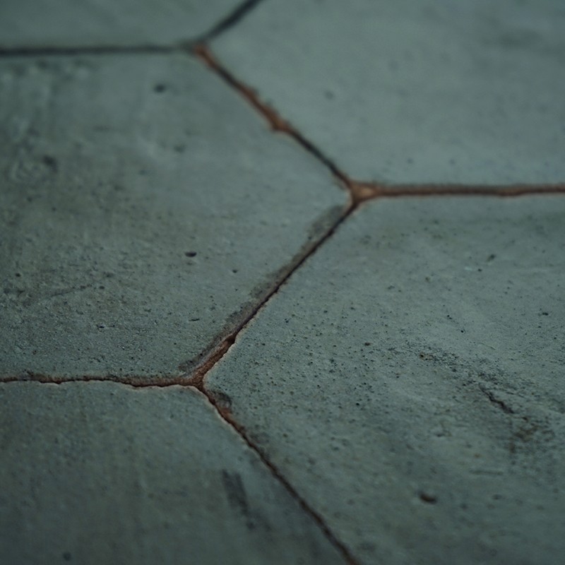 Azulejo hexagonal verde-azulado agrietado y rugoso; juntas marrón rojizas con suciedad, superficie moteada y pequeñas erosiones, aspecto de suelo o pavimento visto de cerca.