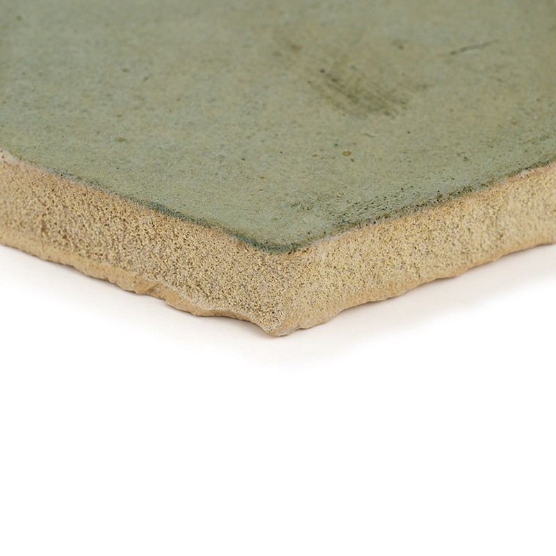 Azulejo cerâmico — canto com esmalte verde levemente gasto sobre corpo de cerâmica porosa bege, apoiado sobre fundo branco.