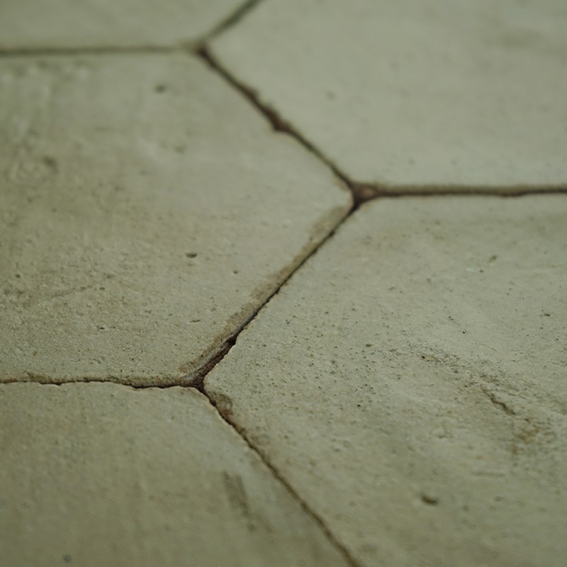 Azulejo hexagonal cerámico envejecido con grietas y juntas oscuras; rodeado por azulejos adyacentes formando una superficie rugosa y ligeramente verdosa.