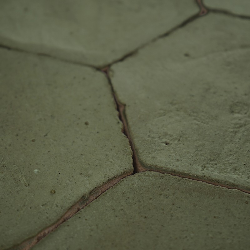 Baldosa hexagonal agrietada; juntas de mortero expuestas y suciedad entre las piezas, tono verdoso apagado, pavimento exterior irregular.