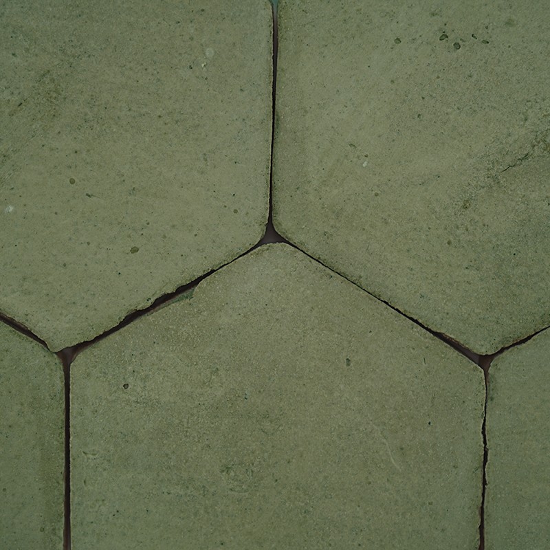 Hexagonale hellgrüne Fliese mit feinen Rissen und dunklen Fugen; enge, leicht sandige Oberfläche, Teil eines regelmäßigen Bodenpflasters.