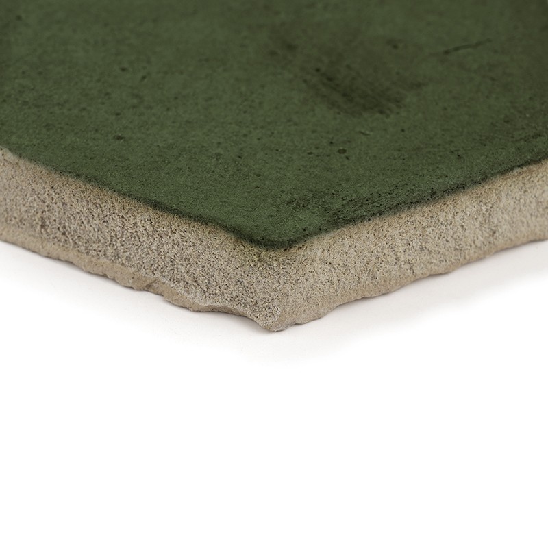 Groene geglazuurde keramische tegel met licht afgeronde hoek; onregelmatige beige poreuze onderzijde zichtbaar, geplaatst tegen een gladde witte achtergrond.