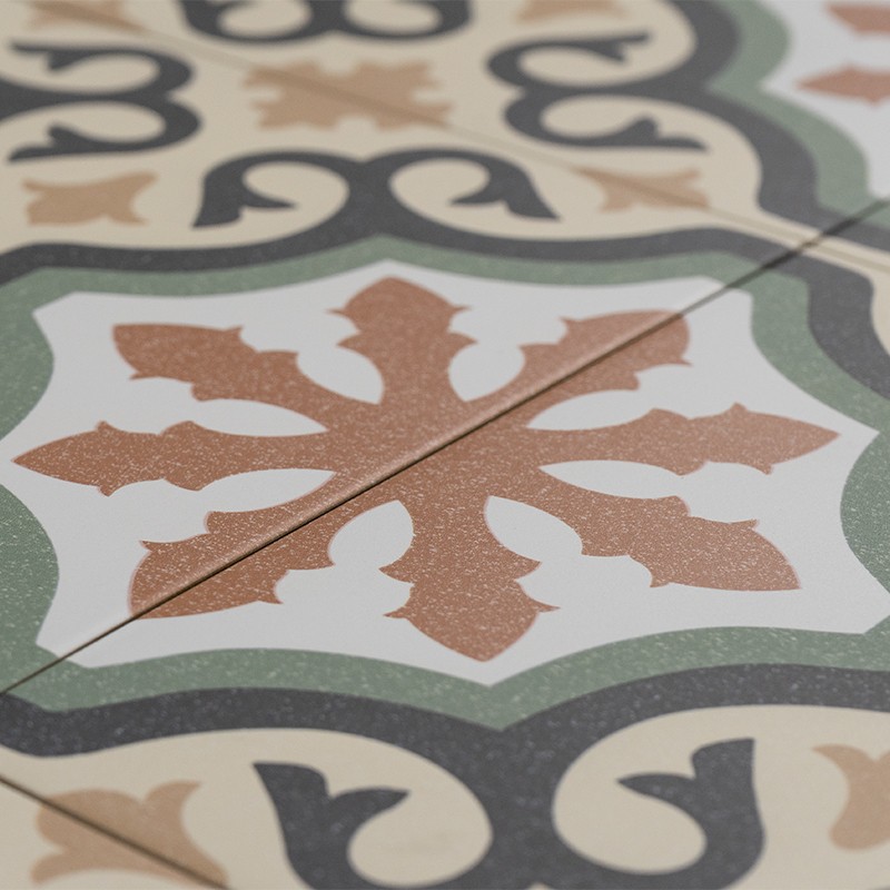 Carreau décoratif à rosace centrale ocre sur fond blanc, bordure verte et arabesques noires; plusieurs carreaux assemblés au sol avec joints brun clair.