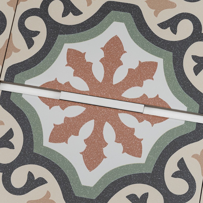 Carreau céramique décoratif: motif floral ocre au centre, fond blanc entouré d'anneaux verts et arabesques noires, joint fin et bande plastique blanche diagonale sur sol carrelé.