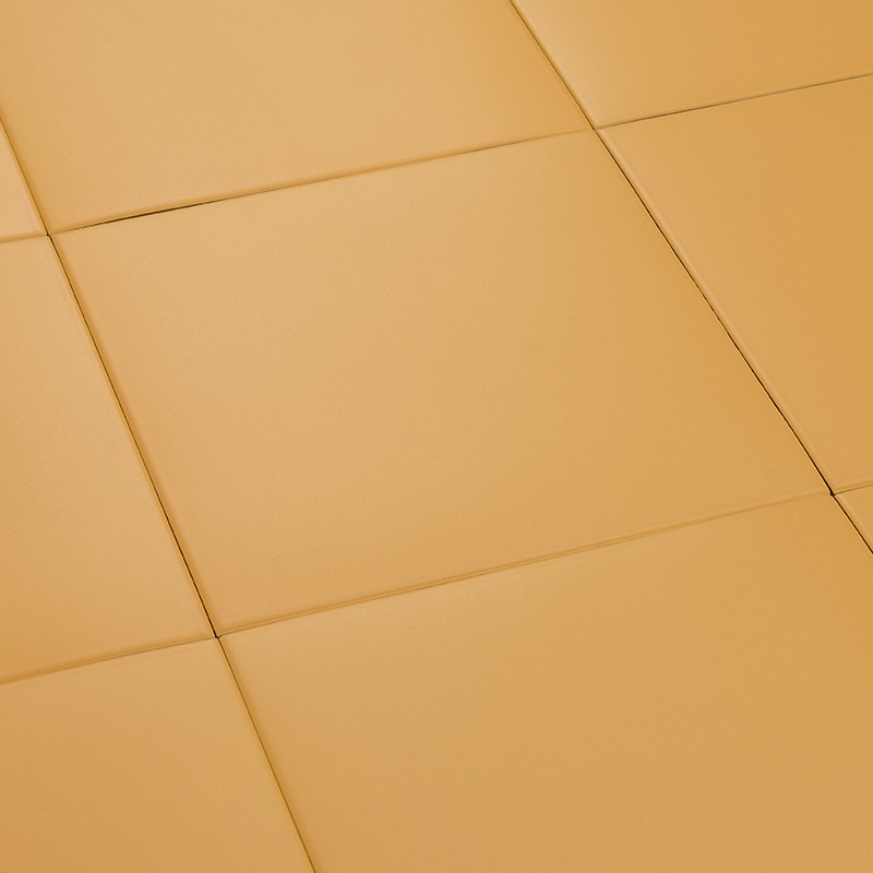 Azulejo cuadrado liso color mostaza; paneles contiguos forman un pavimento uniforme con juntas finas y acabado mate, iluminado por luz cálida.
