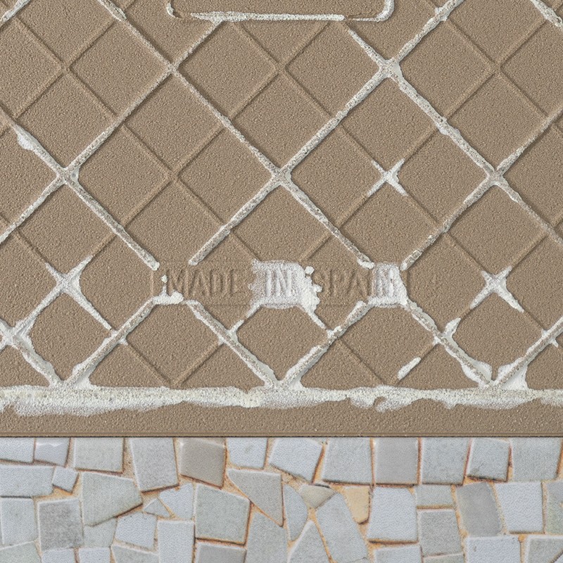 Baldosa cerámica beige con patrón romboidal y texto en relieve "MADE IN SPAIN"; contexto: borde inferior muestra mosaico de pequeños azulejos blancos con lechada.