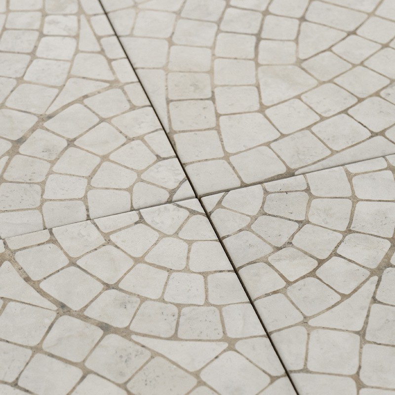 Azulejo cerámico con patrón de pequeños mosaicos formando arcos concéntricos; juntas visibles crean una línea diagonal, superficie de piso en tonos crema con lechada gris.