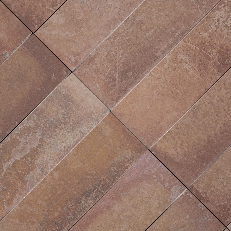 Carrelage rectangulaire brun-rouge disposé en diagonale, surface patinée et veinée, joints fins sombres, couvrant un sol intérieur ou une terrasse.