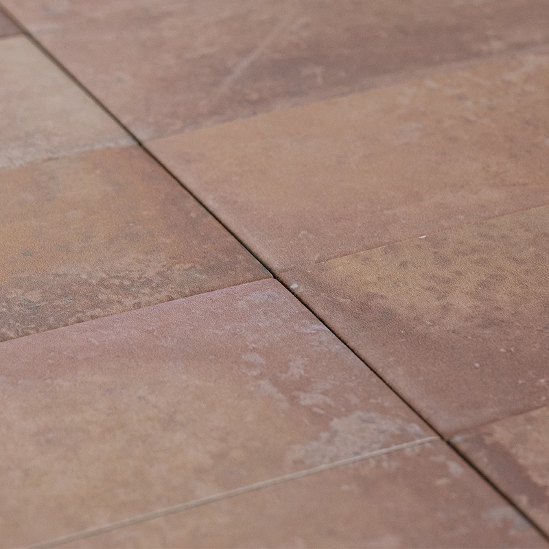 Carrelage en carreaux terre cuite, joints fins foncés; surface légèrement érodée et tachetée, gros plan oblique montrant deux dalles et une jonction diagonale.