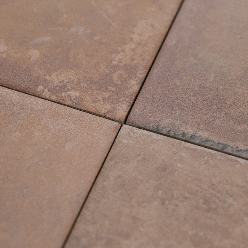 Four brown ceramic tiles arranged in a cross; narrow joints with dirt, slightly worn texture, mottled tones and beveled edges.