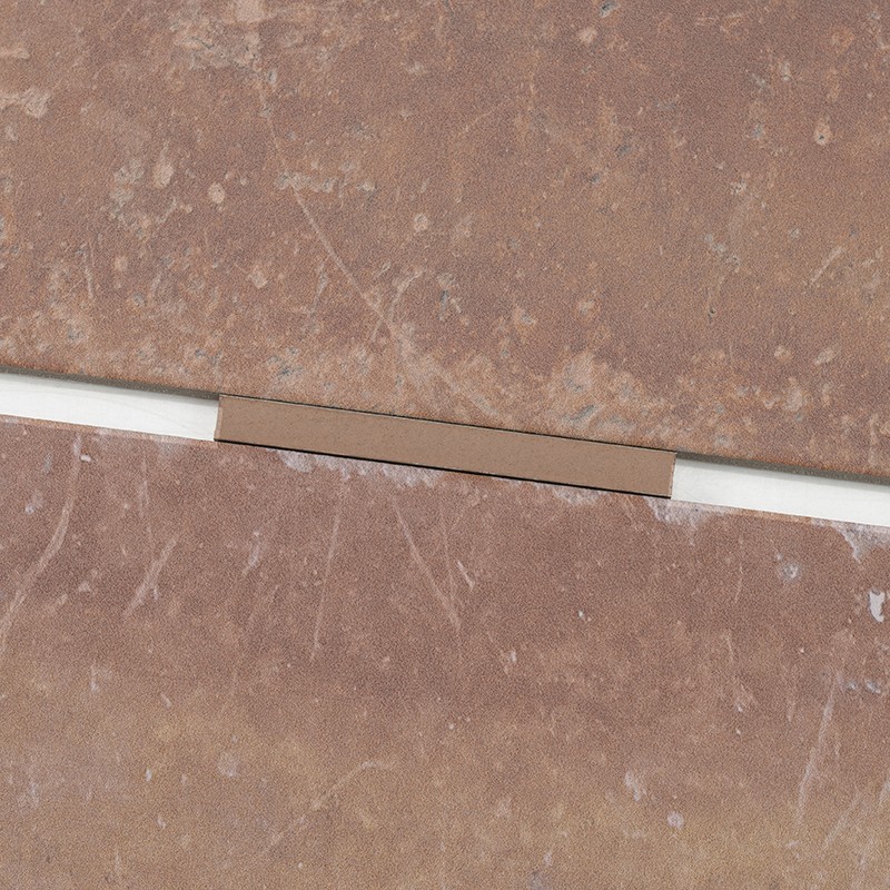 Reddish-brown tile; a white joint between pieces, partially covered by a small brown rectangle; textured surface with speckles and stripes.