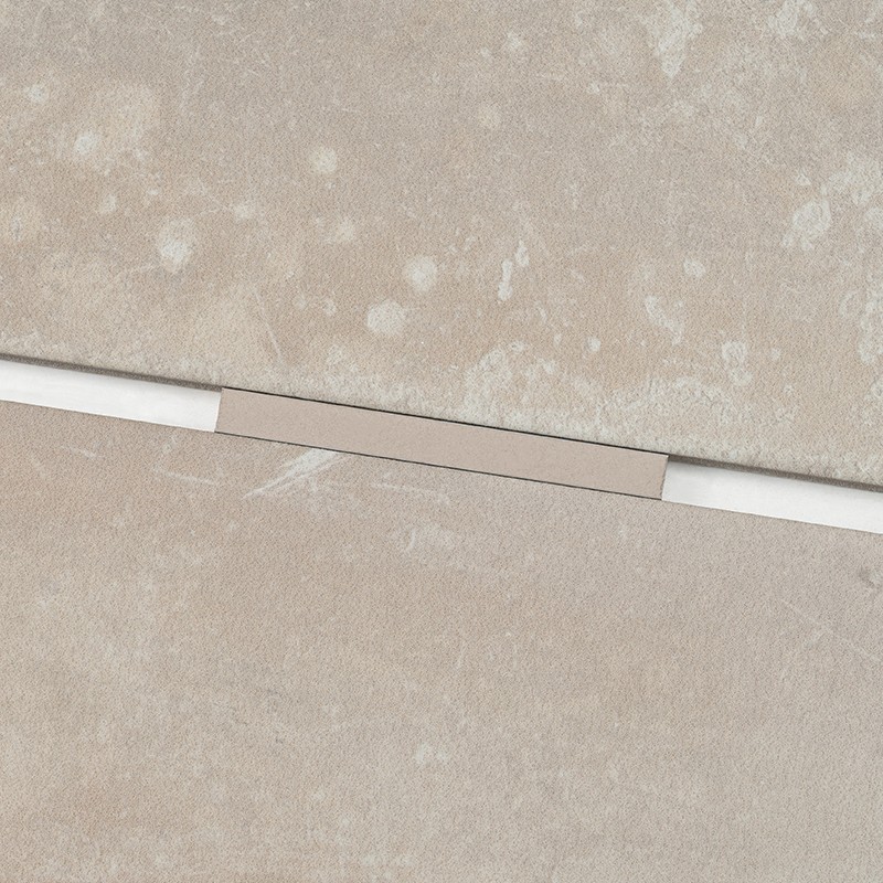 Azulejo beige marmoleado rectangular; junta horizontal blanca con tira metálica estrecha encajada en el centro; azulejos contiguos del mismo tono.