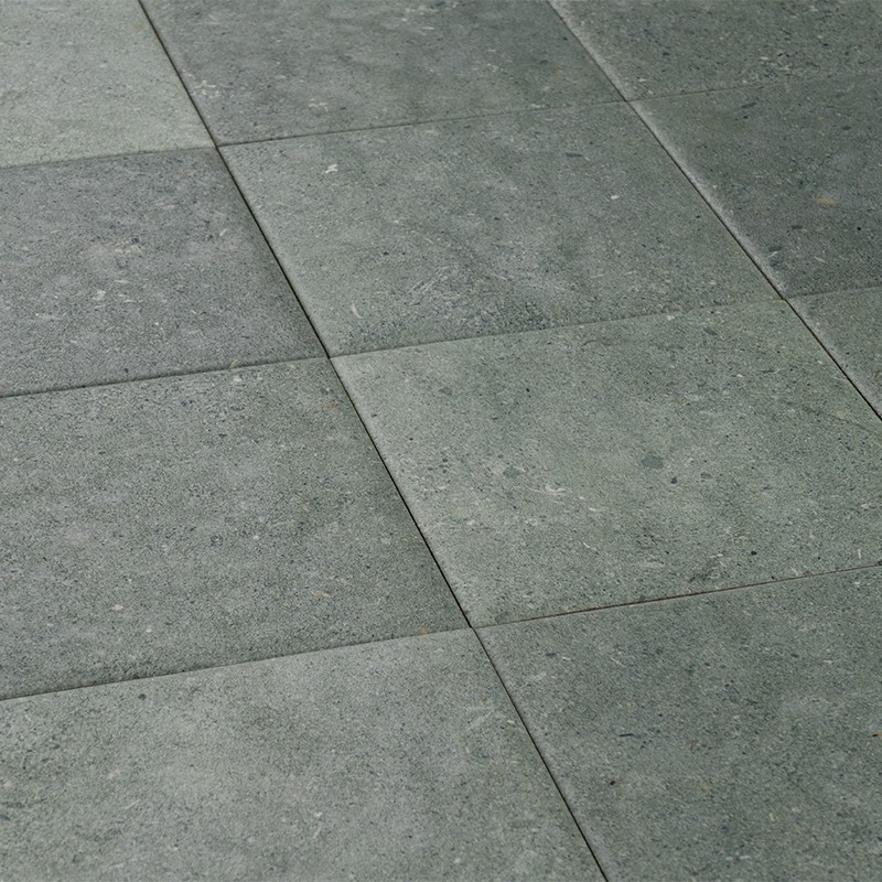 Vierkante grijsgroene vloertegels met subtiele textuur en lichte vlekken, netjes geplaatst in een raster met dunne voegen; schuin perspectief toont meerdere aangrenzende rijen.