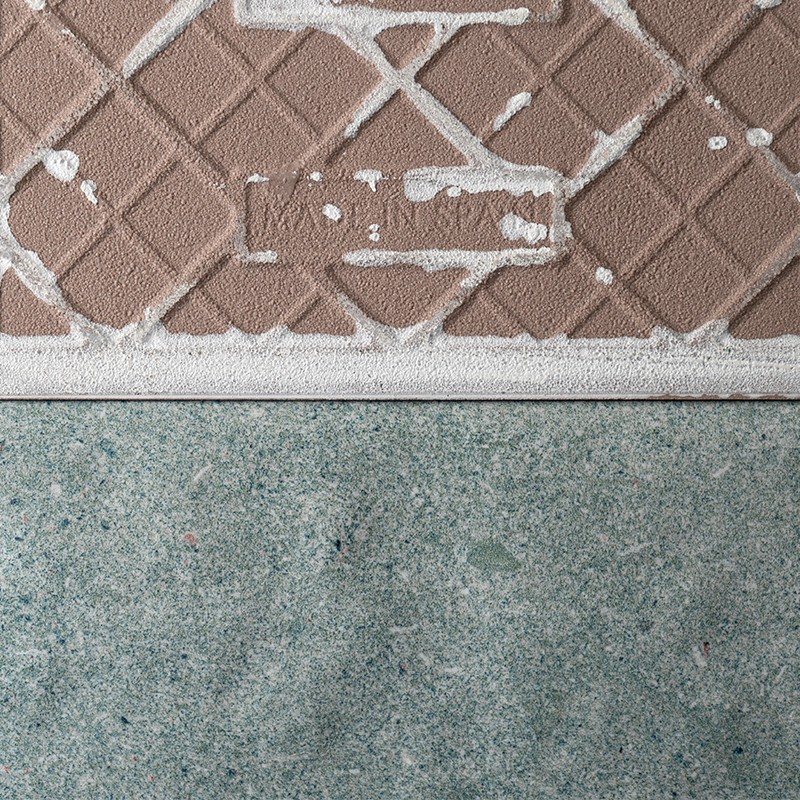 Carrelage brun à motifs losanges, éclaboussures et coulures de peinture blanche, séparé par une bande claire d'un sol en béton vert‑gris.