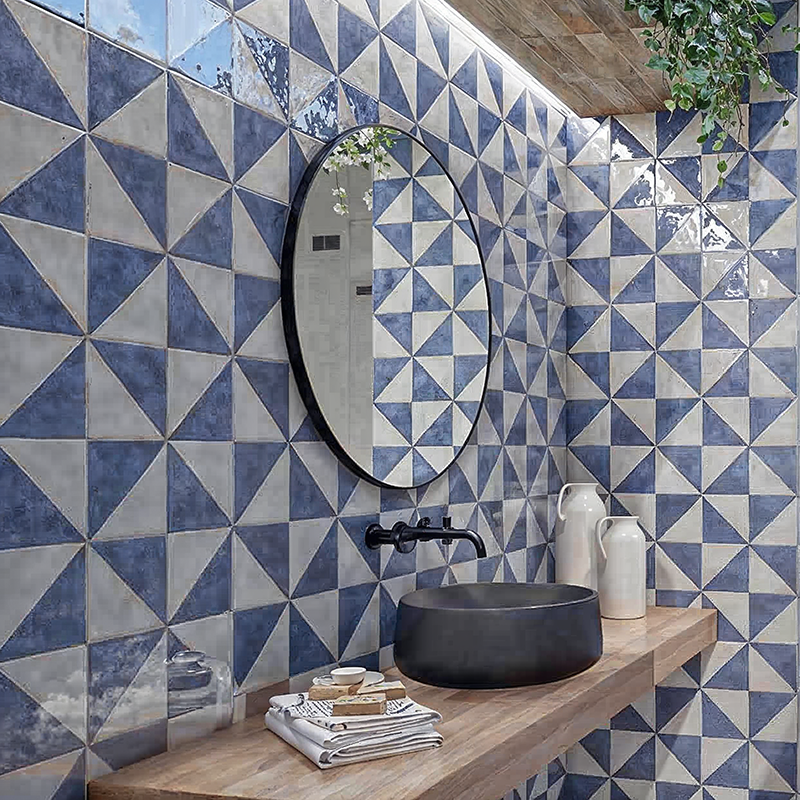 Blau-weißes, geometrisches Fliesenmuster (Dreiecke) an Wänden; runder Spiegel, schwarzes Wandbecken und Armatur auf Holzablage, weiße Keramikgefäße und hängende Pflanze.