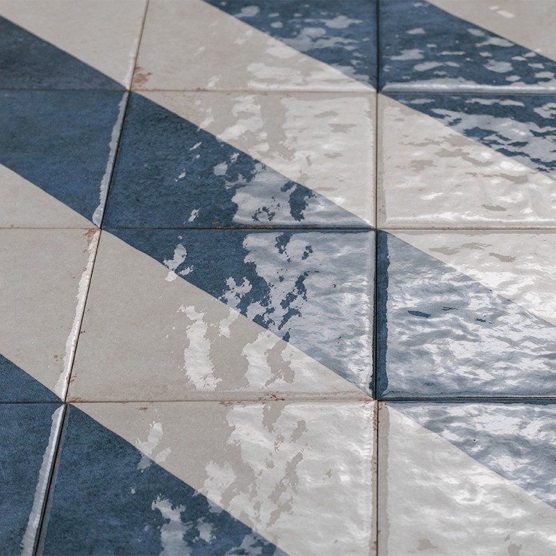 Carrelage en carreaux carrés bleus et blancs formant un motif diagonal de triangles, finition brillante avec reflets, joints beiges visibles et légère usure sur certaines surfaces.