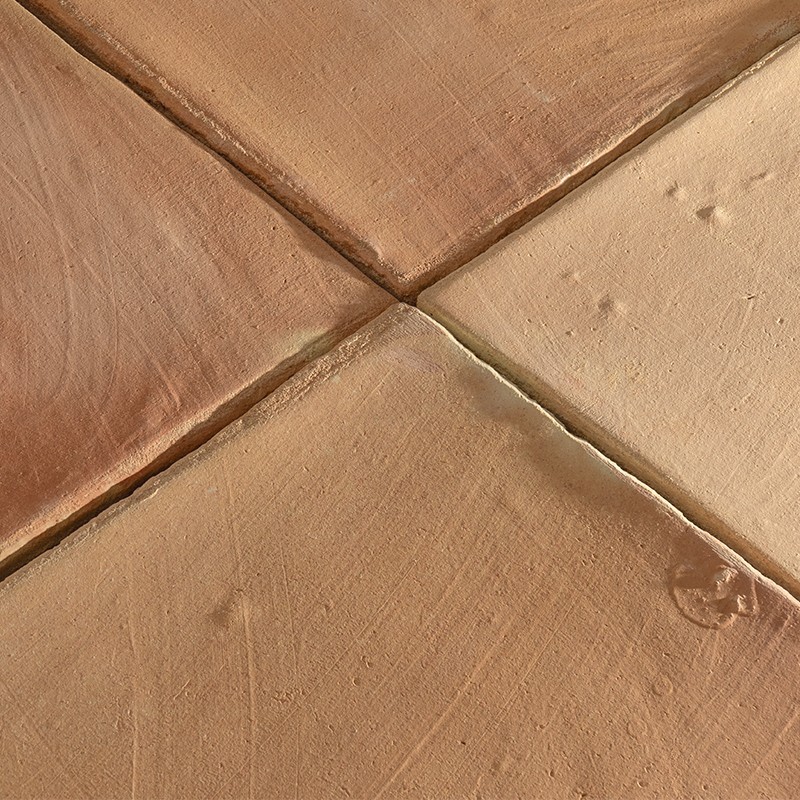 Azulejo de terracota, superficie mate con pequeñas hendiduras y marca circular; juntas en cruz entre cuatro piezas; contexto: suelo rústico de interior.