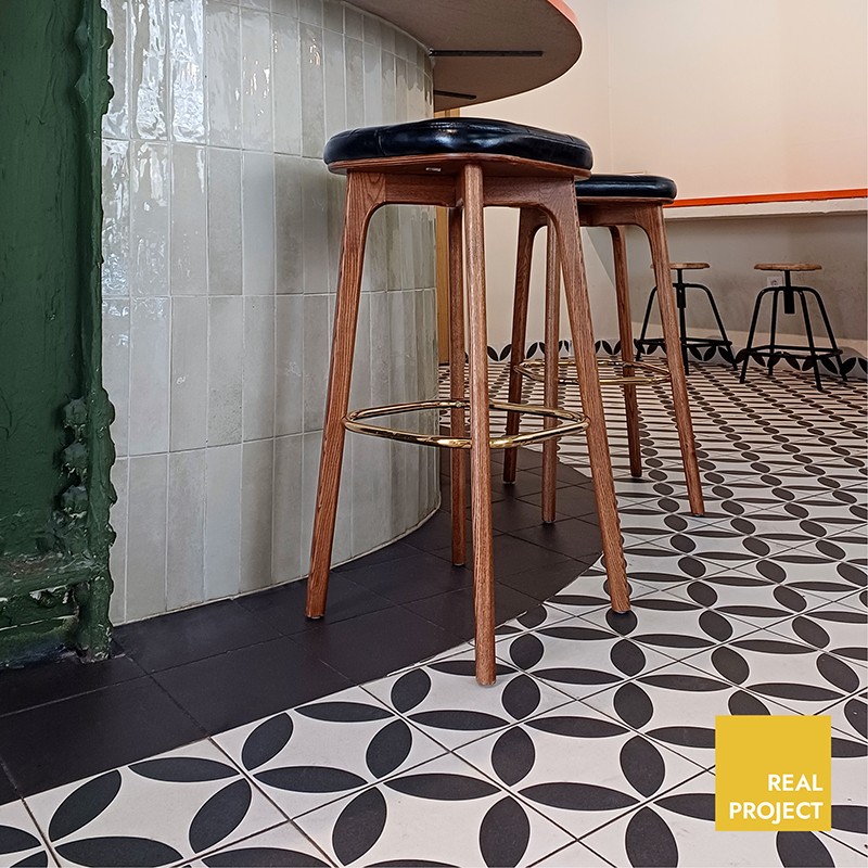 Carrelage noir et blanc à motifs géométriques au premier plan; tabourets en bois et cuir devant un comptoir carrelé incurvé, intérieur de café/restaurant.  Texte visible : "REAL PROJECT"