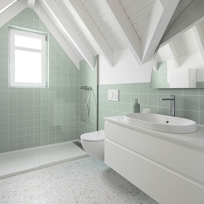 Azulejo verde-menta em banheiro sob sótão com vigas brancas, box de piso aberto, chuveiro de parede, vaso suspenso, bancada e lavatório brancos, piso claro pontilhado.
